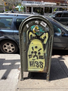 A yellow, angry looking cat is graffiti-ed on a mailbox on a sidewalk with the word "Hiss" written across the bottom.  
